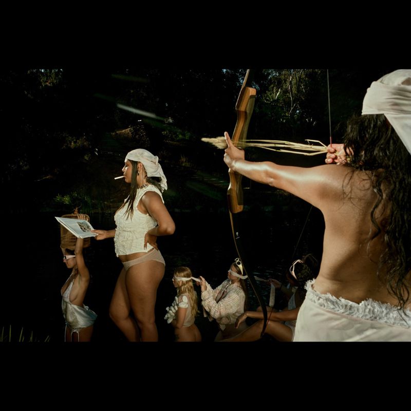 Group of women dressed in white fabrics. One woman draws back a wooden bow. One stands to the side holding a cigarette and reading a magazine.