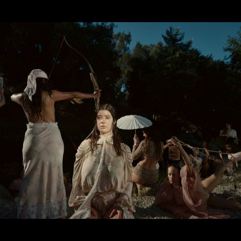 Group of women in ethereal dresses pose with a bow and arrow in a dark outdoor setting with trees, creating a surreal and artistic composition.