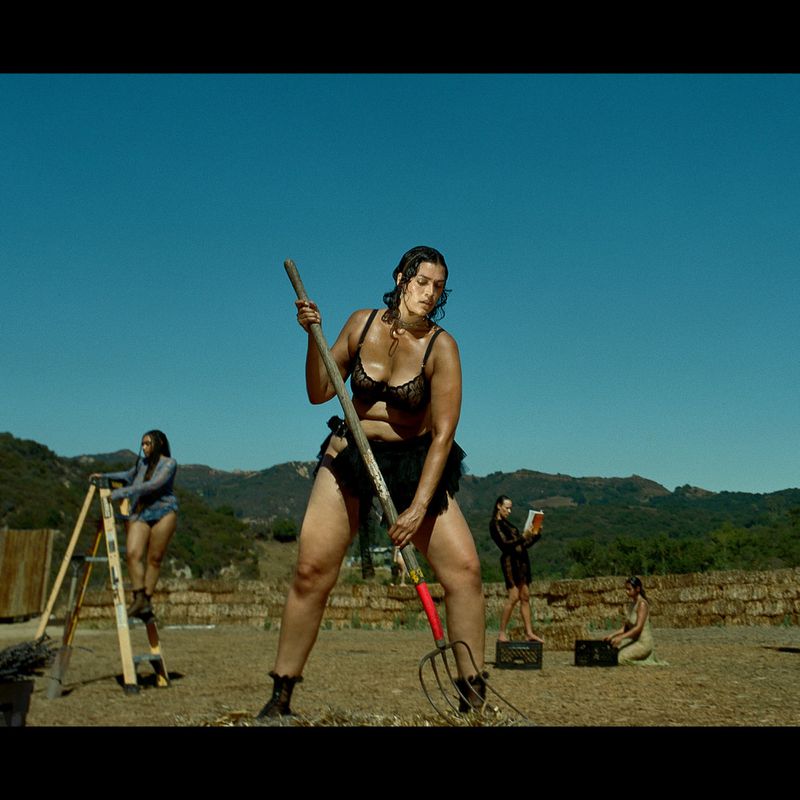 Woman in lingerie stands with pitchfork on a farm under a clear sky, with other women and farm buildings in the background.