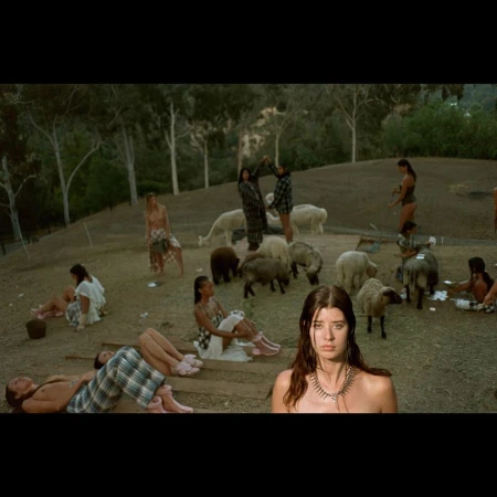 Outdoor gathering with a group of people and sheep in a field, featuring an adult woman in the foreground and lush green trees.