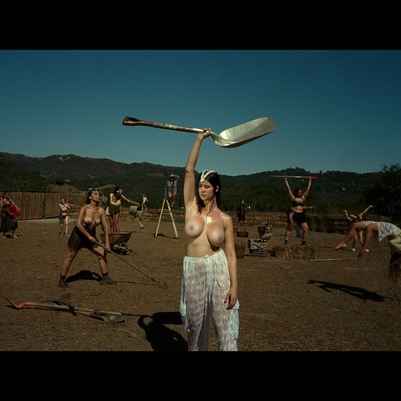 Group of partially nude women working in a field, one holding an oversized spoon overhead, under a clear blue sky.
