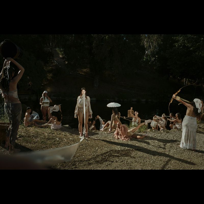 Group of women in outdoor setting, one pouring water from bucket, another with bow and arrow; trees and water in background.