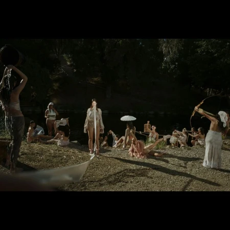 Group of women in outdoor setting, one pouring water from bucket, another with bow and arrow; trees and water in background.
