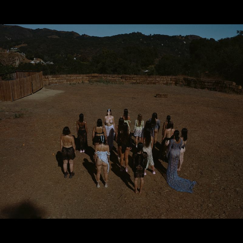 Group of people standing outdoors in a circle formation on dry land, under dark lighting, mountains and a wooden fence in the background.