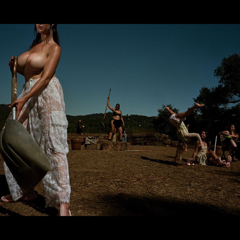 Nude woman in a lace skirt holds a shovel in an outdoor field, with multiple people in the background under a clear blue sky.