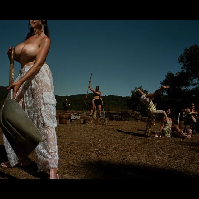 Nude woman in a lace skirt holds a shovel in an outdoor field, with multiple people in the background under a clear blue sky.