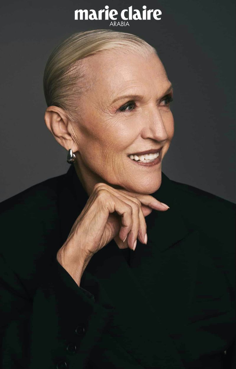 Close-up portrait of an older woman with short blonde hair, wearing a black blazer and silver hoop earrings against a grey backdrop.