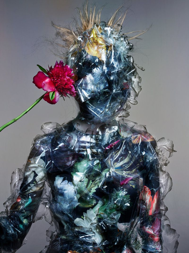 Artistic portrait of person in floral bodysuit, covered with plastic, flower by face, against grey backdrop. Unusual, creative fashion.