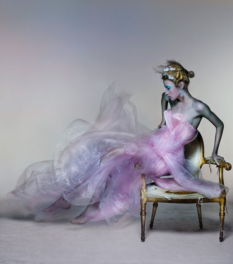 Young woman with hair up and fantasy makeup sits on a gold antique chair in front of a neutral background with a flowing sheer gown.