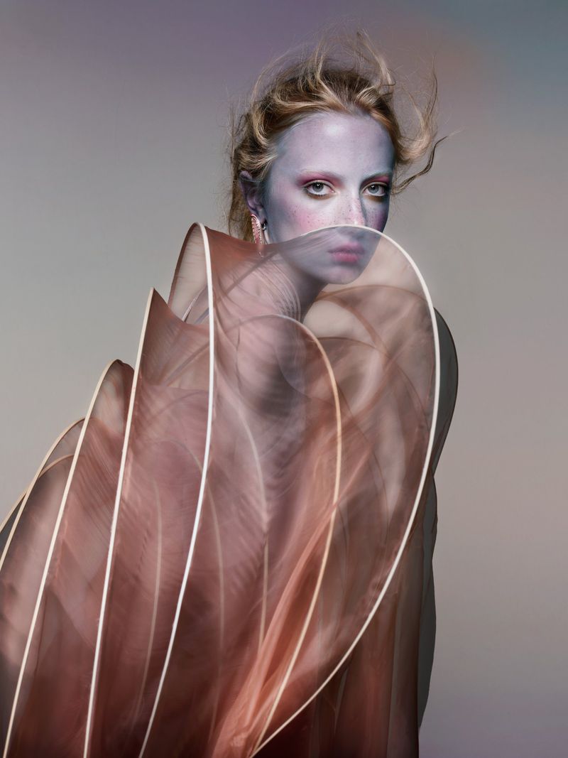 Eye-level portrait of a blond adult female with dramatic makeup wearing a sheer, brown, flowing garment against a neutral gradient background.