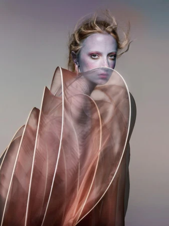 Eye-level portrait of a blond adult female with dramatic makeup wearing a sheer, brown, flowing garment against a neutral gradient background.