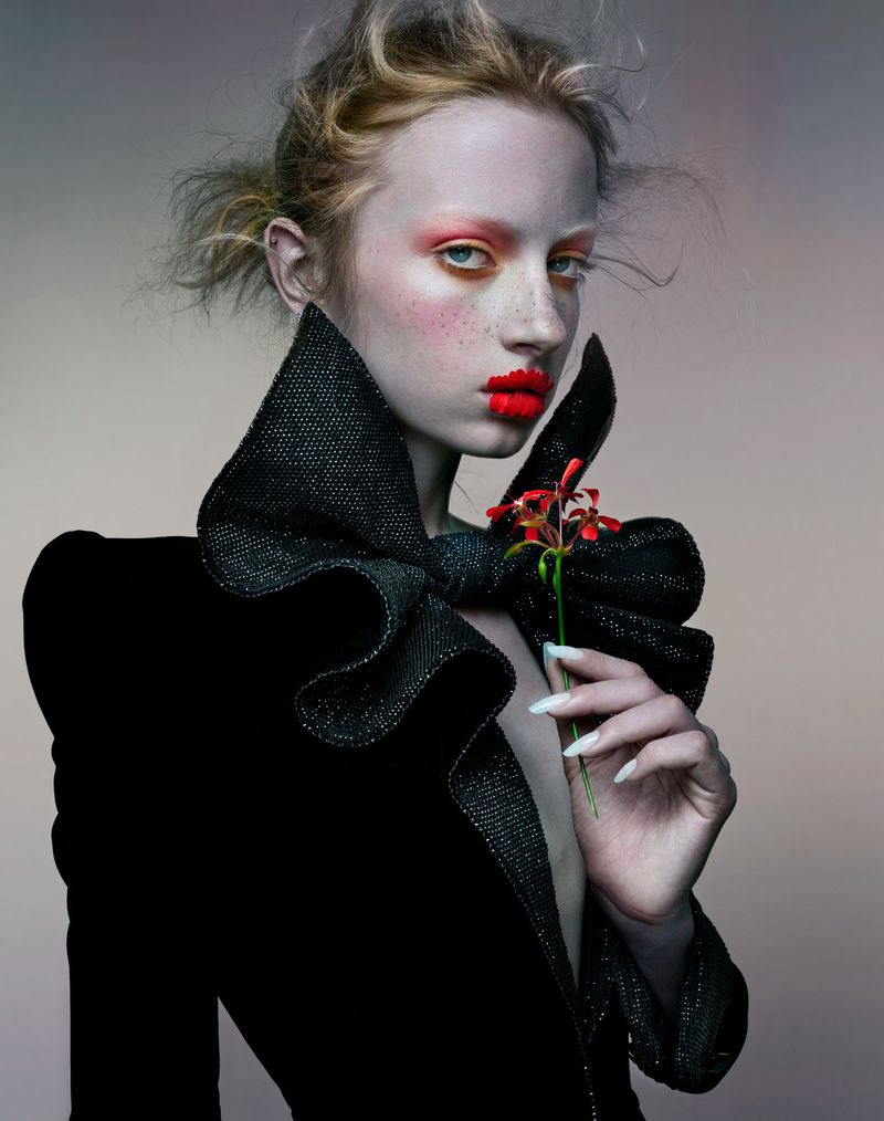 Woman with pale skin and bold makeup in a black textured jacket holding a red flower, in a fashion-forward studio portrait.