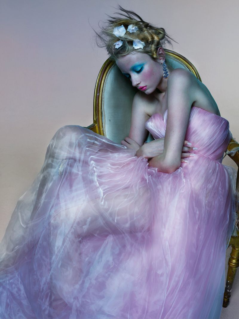 A young woman with blonde hair and vibrant blue makeup is dressed in a pink tulle gown, seated in an ornate chair, in a fashion editorial.