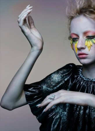 Young woman with unique artistic makeup and a shiny black top poses in front of a soft, neutral backdrop with studio lighting.