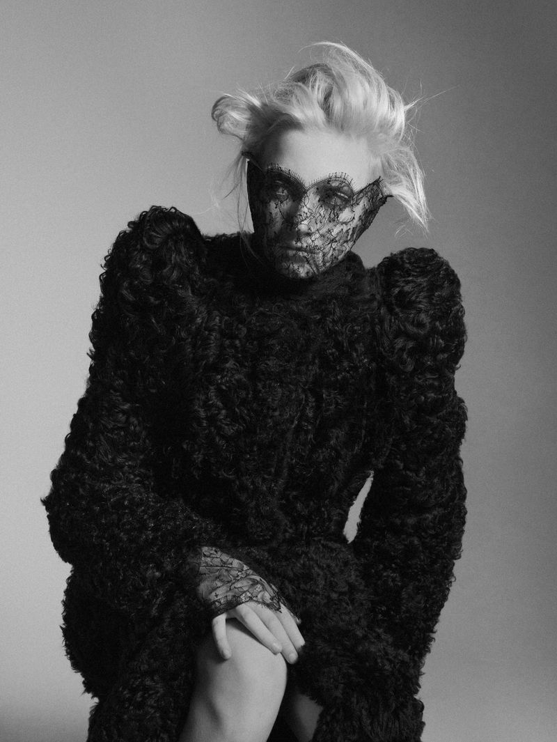 Woman in a textured black coat with lace detail on face and hands, monochromatic portrait in front of a neutral background.