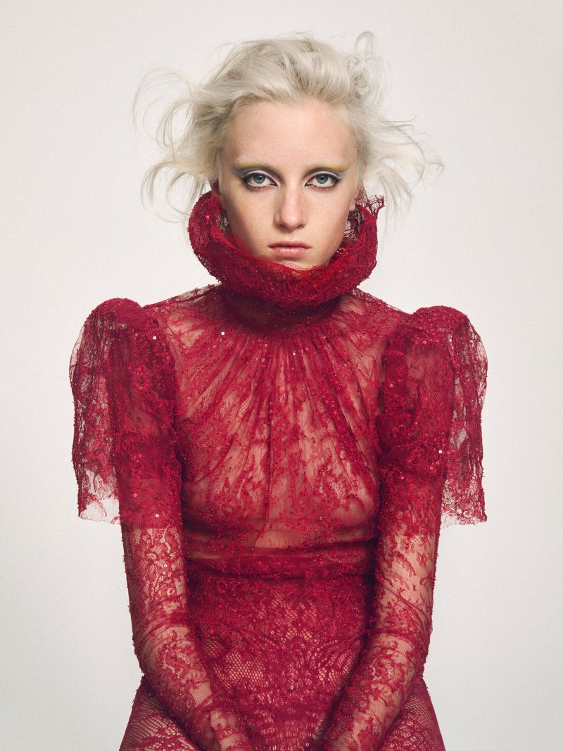 A fair-skinned woman with platinum blonde hair and yellow eye shadow wears a red, lace, long-sleeved dress with a high ruffled collar against a white backdrop.