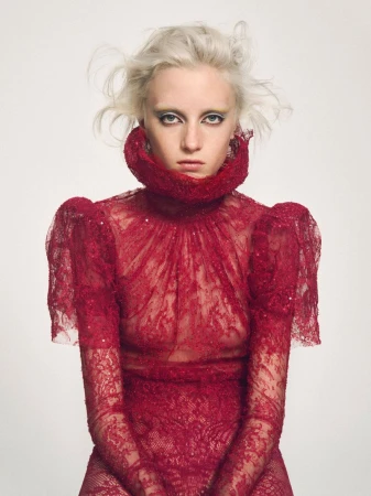 A fair-skinned woman with platinum blonde hair and yellow eye shadow wears a red, lace, long-sleeved dress with a high ruffled collar against a white backdrop.
