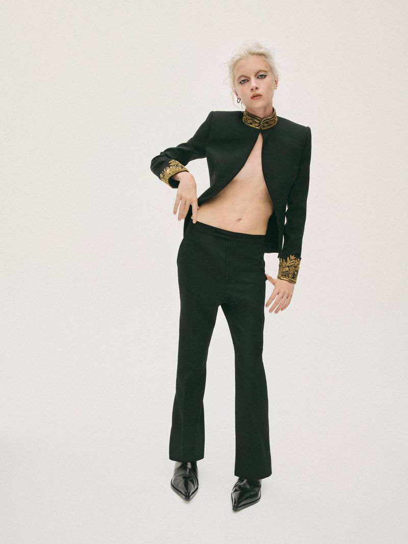 A fair-skinned adult woman with short blonde hair poses in a black suit with gold trim against a white backdrop in a studio setting.