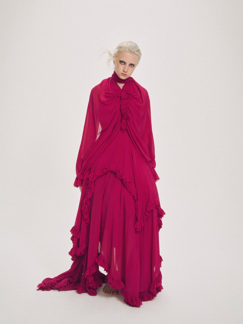 A light-skinned woman with blonde hair in a floor-length magenta gown poses in front of a white backdrop in a fashion forward portrait.