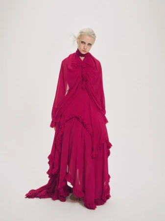 A light-skinned woman with blonde hair in a floor-length magenta gown poses in front of a white backdrop in a fashion forward portrait.