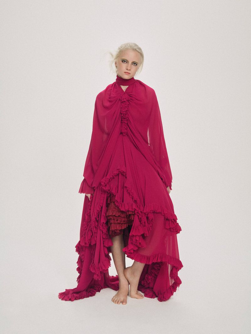 Full-length portrait of a young adult female with blonde hair and fair skin, wearing a ruffled, long-sleeved, magenta gown on a white backdrop.