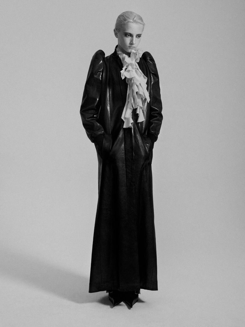 Monochromatic studio shot of a fair-skinned adult woman with platinum blonde hair posing in a shiny, black, full-length leather coat with ruffled collar.