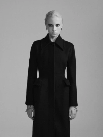 Model in black coat wearing earrings and bracelets against a grey seamless background for Beyond Noise by Valeria Herklotz