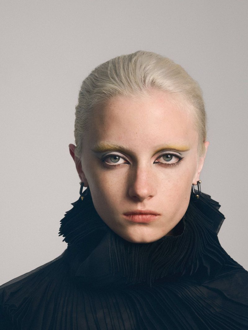 Close-up portrait of a young woman with short blonde hair and dramatic yellow eye makeup, wearing a textured black top against a plain background.