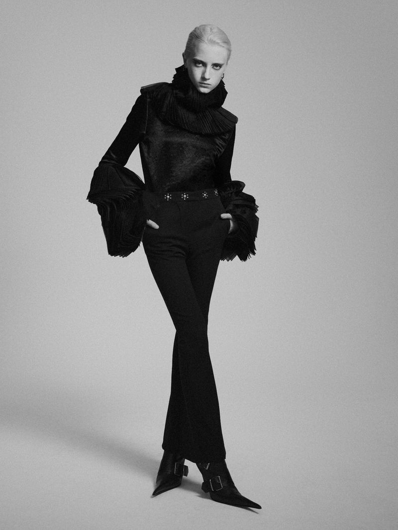 Full-length black and white fashion portrait of a woman with short white hair wearing a black ruffled collar shirt and pants in a studio.