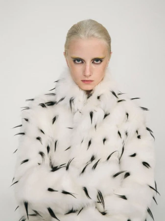 A fair-skinned woman with light blonde hair stares intently at the camera while wearing a white fur coat with black accents.