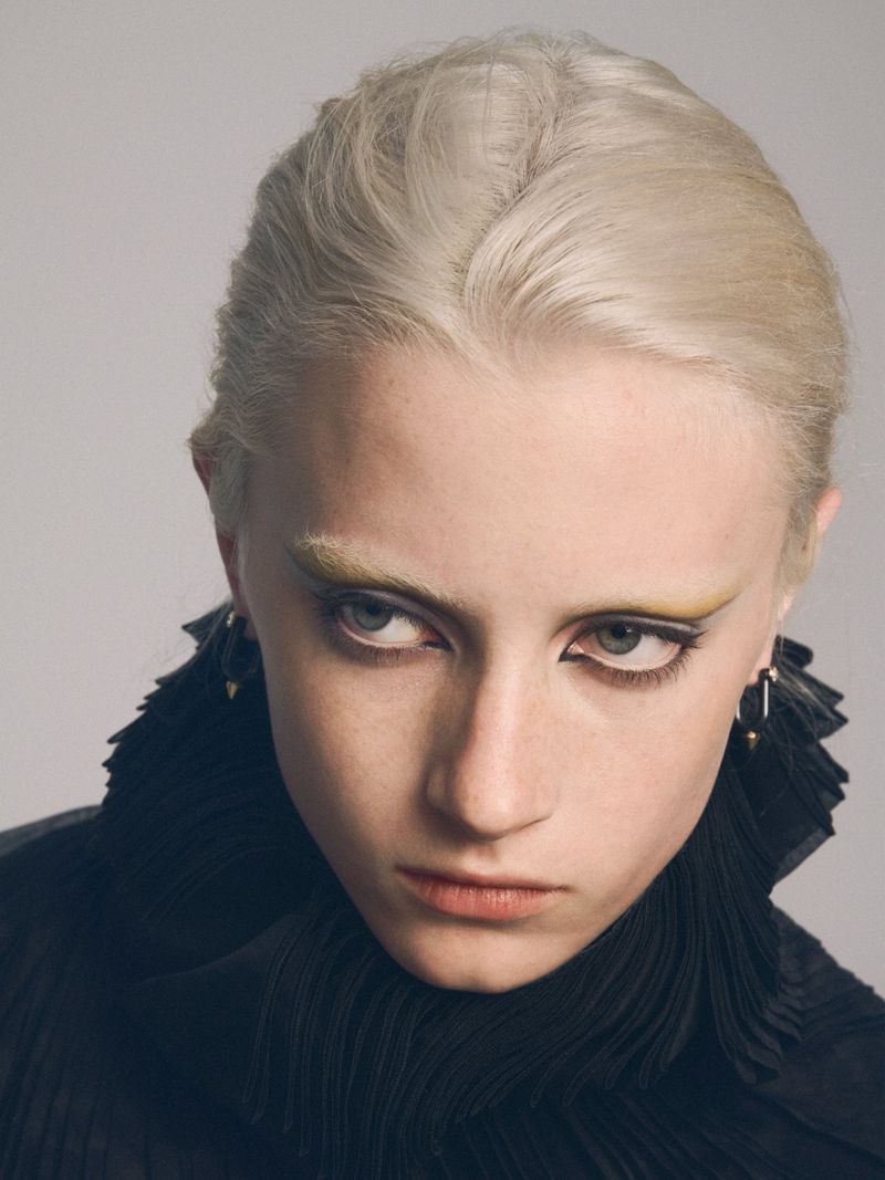 Close-up studio shot of a fair-skinned adult female model with platinum blonde hair, dramatic yellow makeup, wearing a black ruffled collar.