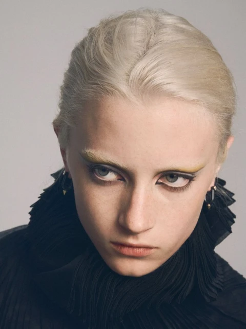 Close-up studio shot of a fair-skinned adult female model with platinum blonde hair, dramatic yellow makeup, wearing a black ruffled collar.