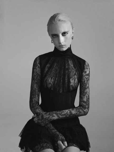 Monochrome portrait of a fair-skinned adult woman with blonde hair wearing a black lace dress against a plain grey background.