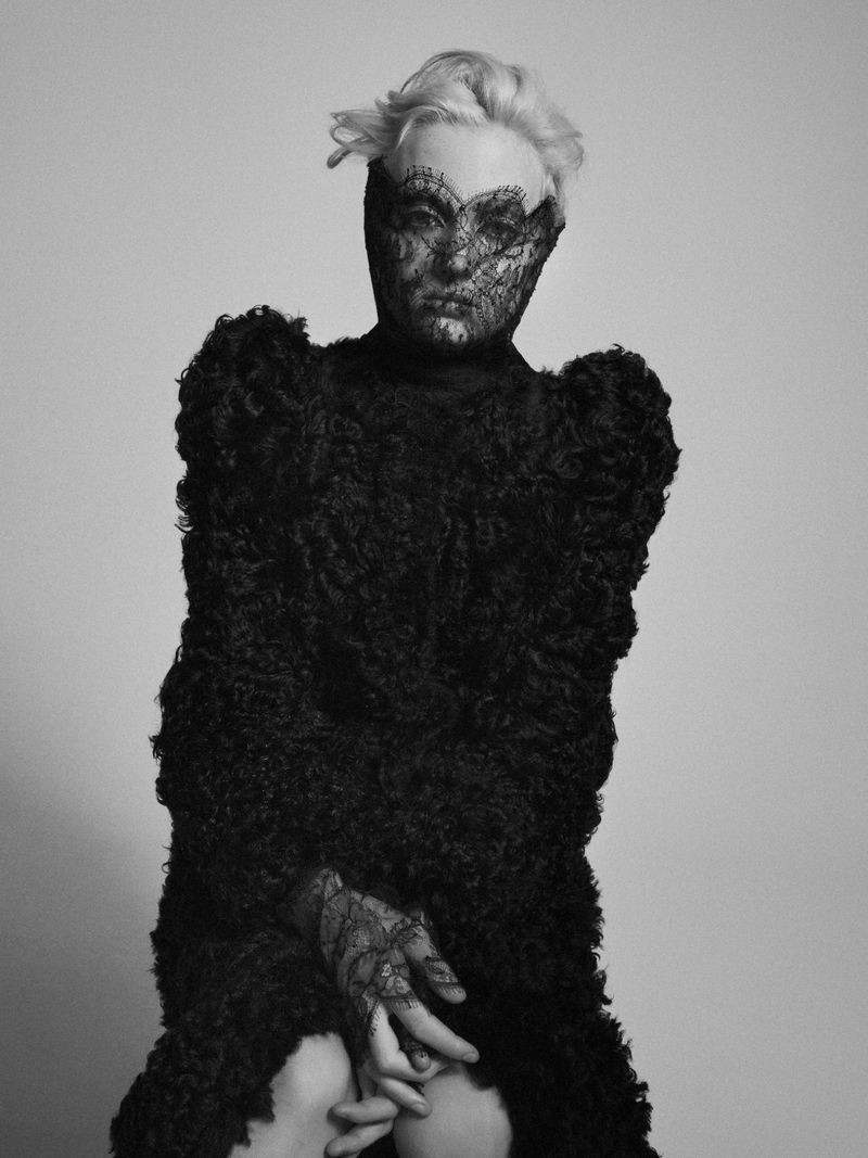 Striking black and white portrait of a stylish adult woman with short blonde hair, wearing a lacy mask and textured black jacket in a studio setting.