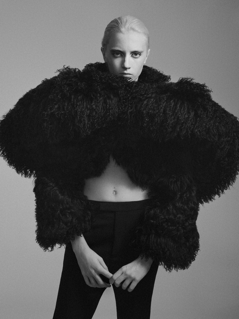 Model Kai Schreiber wearing an oversized textured black coat and dark trousers against a grey backdrop.