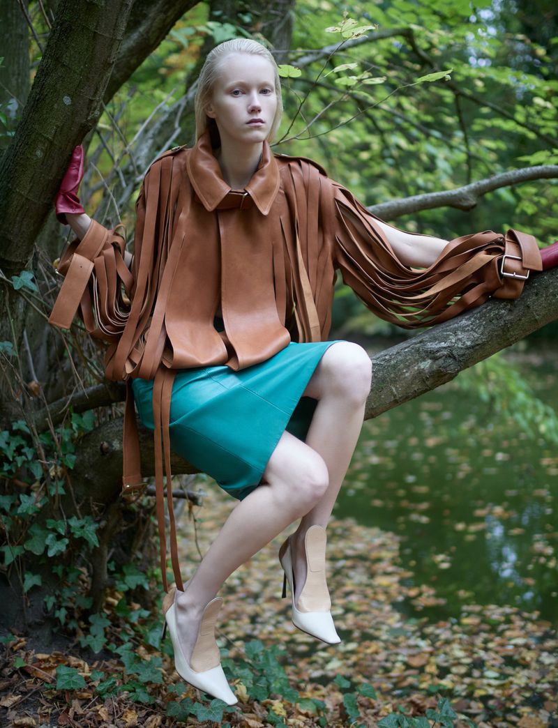 Fashion shot of a model in an unusual brown leather outfit and teal skirt sitting on a tree branch in a forest setting.
