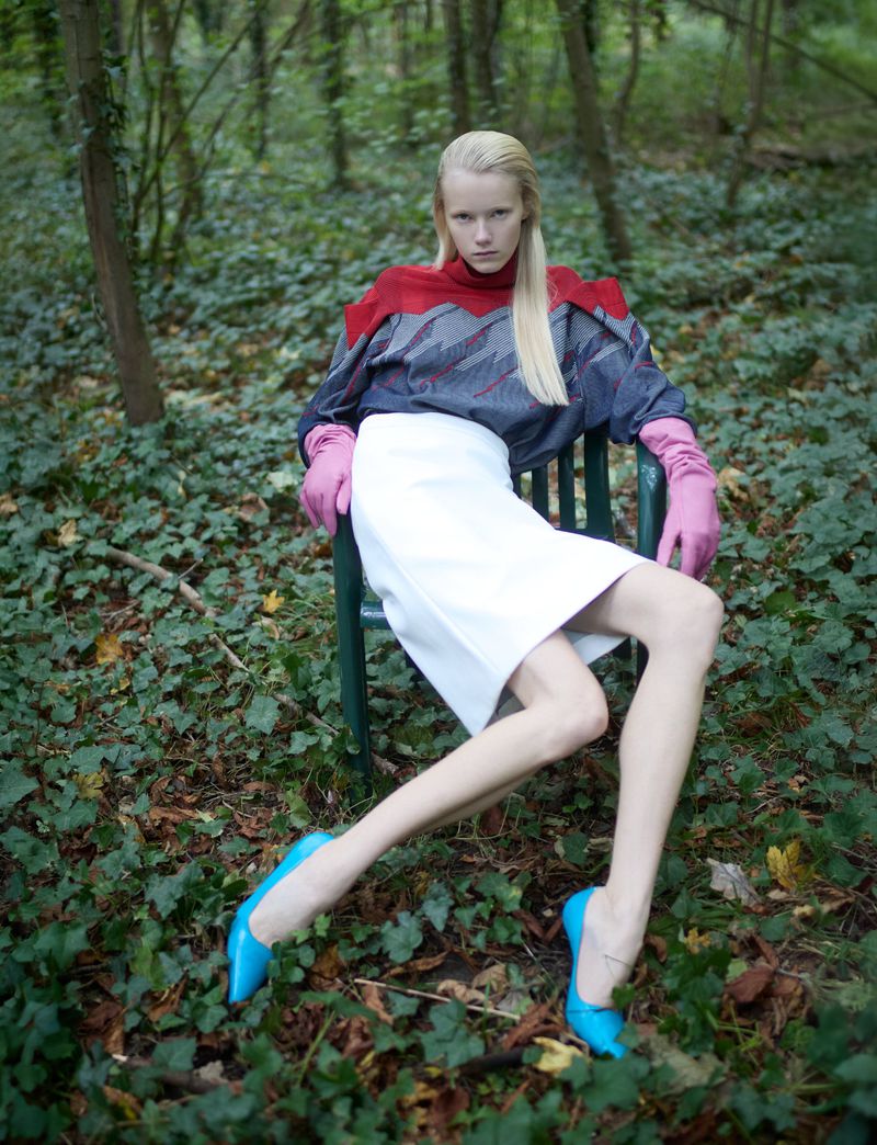 Woman with blonde hair wearing gray top, white skirt, and blue shoes sits in a chair in forest with ivy ground cover.