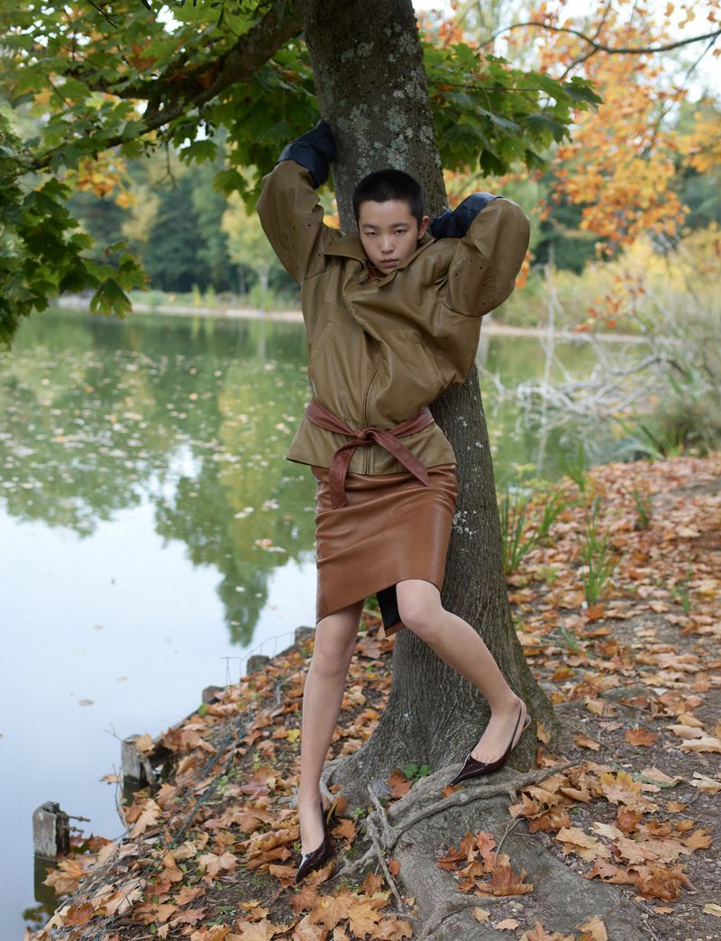 Young woman in brown leather skirt and jacket poses by a tree near a lake with fallen leaves, creating a fashionable outdoor portrait.