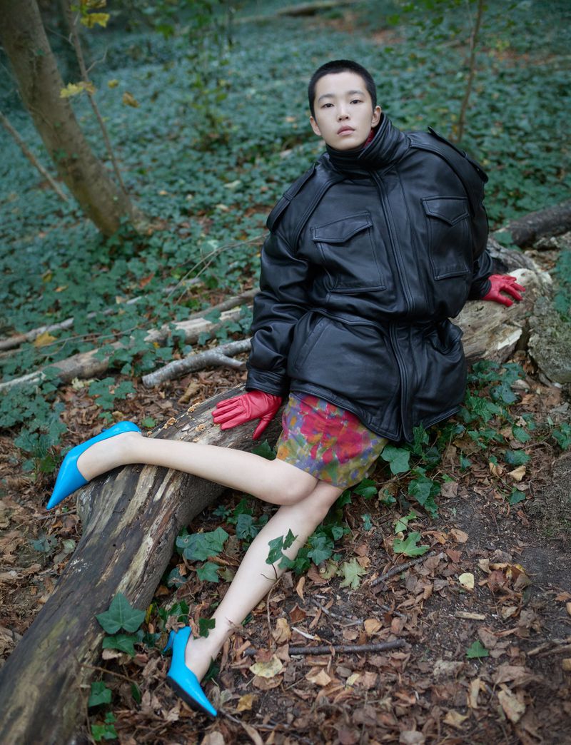 A person in a black jacket, gloves and blue heels lounges on a fallen tree in the woods amid brown leaves and green ivy.