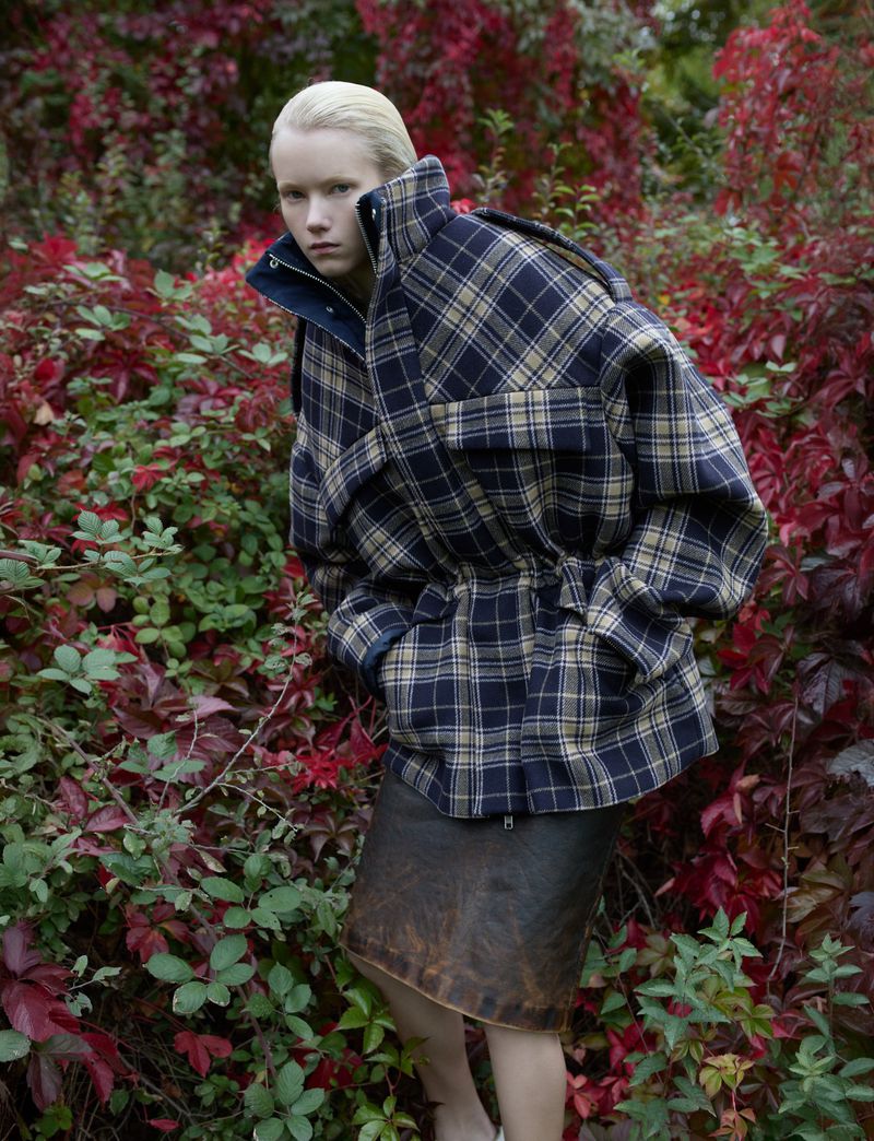 Woman in plaid jacket poses outdoors surrounded by red and green foliage in fashion editorial. Medium shot, eye-level angle, diffused lighting.