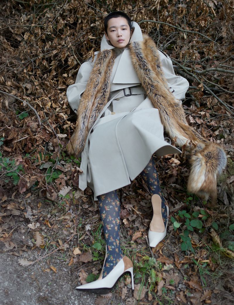 Woman in trench coat with faux fur, patterned tights and heels posing in autumnal forest, fashion editorial lifestyle photography.