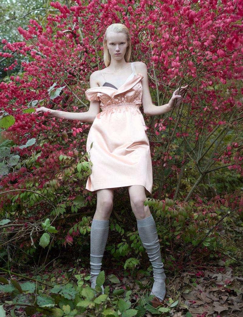 Woman in a pink dress stands among greenery and flowering bushes in an outdoor fashion shot, posing with her arms outstretched.