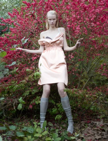 Woman in a pink dress stands among greenery and flowering bushes in an outdoor fashion shot, posing with her arms outstretched.