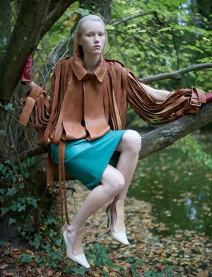Model seated on a tree branch above water wearing a brown fringed jacket and turquoise skirt for Re-Edition Magazine