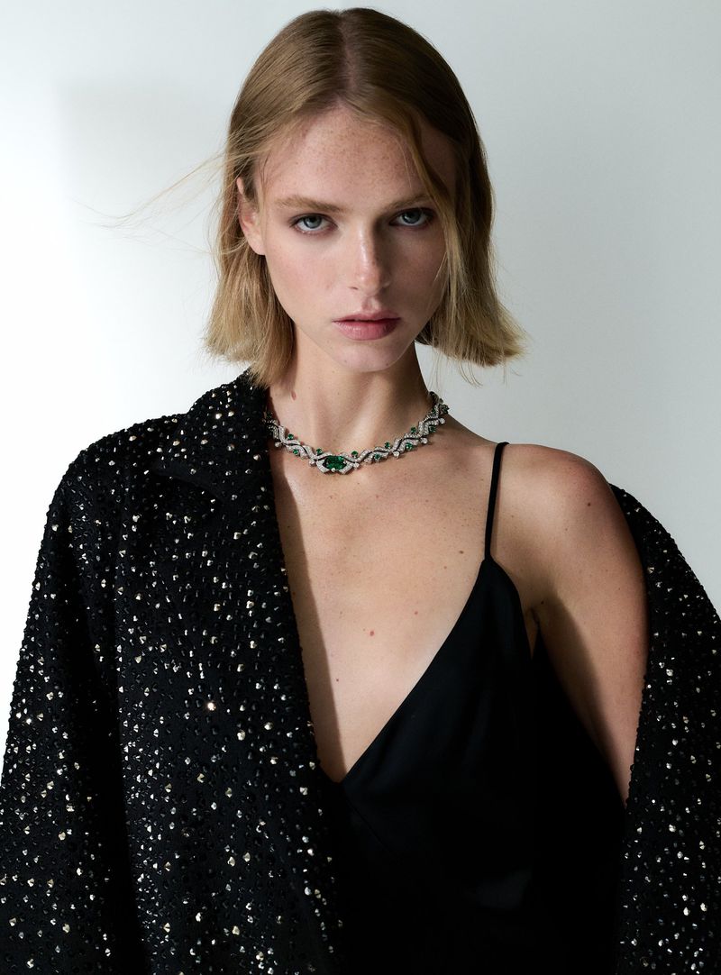 Portrait of a blonde woman wearing an emerald and diamond necklace, with a black sequined jacket against a white backdrop.