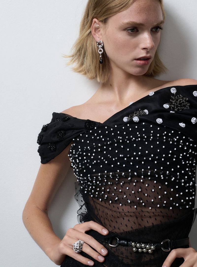 Model in off-the-shoulder black dress with pearl embellishments, posing in a studio. Fashion, portrait, jewelry, and style editorial.