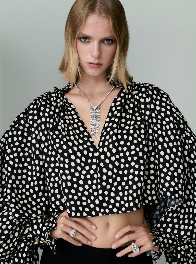 Fashion model poses in a studio wearing a black and white polka dot blouse with jewelry; the portrait features an eye-level angle.