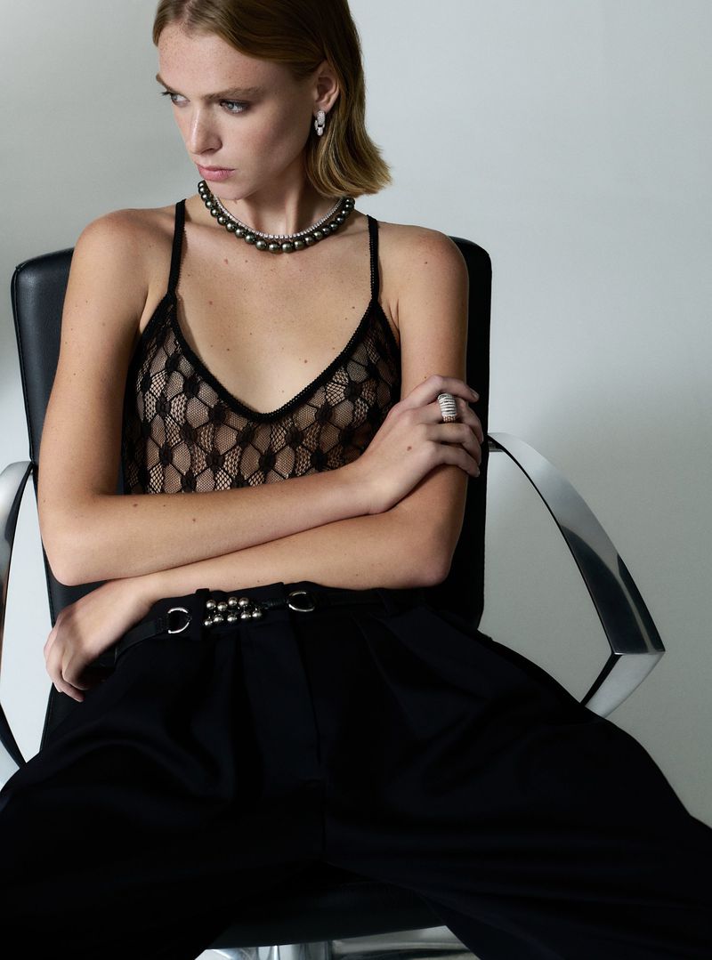 Woman in black lace top and pants poses in a studio setting, wearing pearl necklace and ring, looking off-camera with a neutral expression.