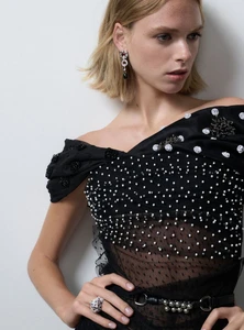 Model wearing a black embellished off-the-shoulder top and crystal earrings against a plain white background