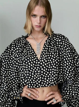 Model in black and white polka dot blouse wearing diamond necklace and rings against grey background